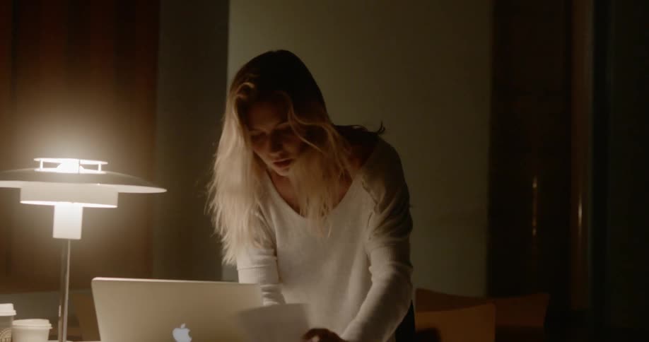 A young woman in a dimly lit room hurriedly looks through her notes on the desk in front of her