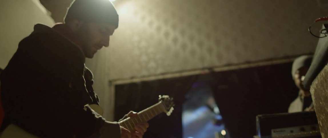 Man plays a guitar as another man moves the audio equipment
