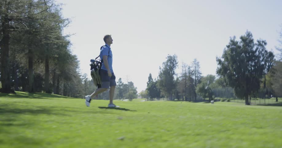 A man walks along a grassy golf course while carrying a large bag of golf clubs over his shoulder