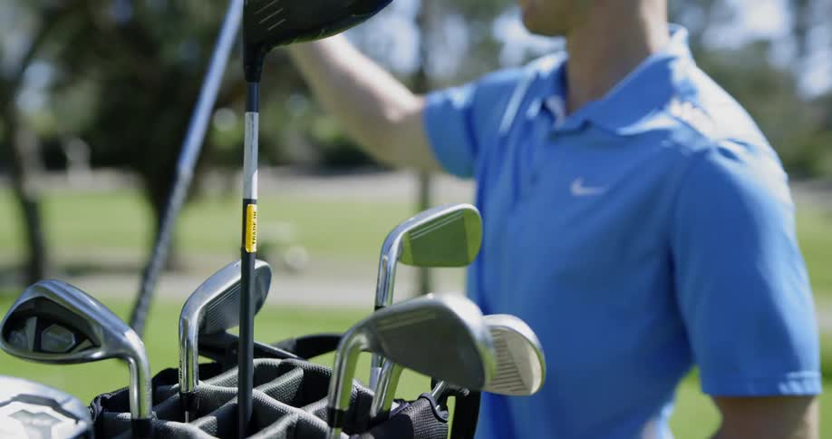A man in a blue polo shirt walks to his golf bag and takes a golf club from it