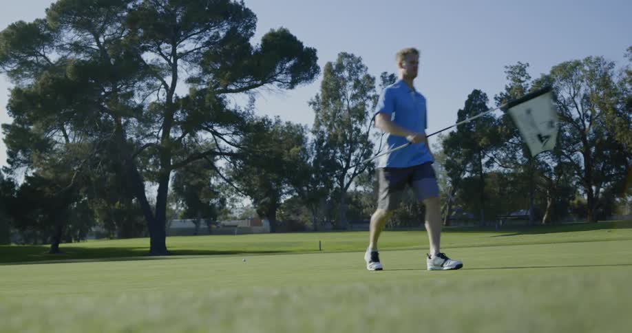 Male carrying a golf club walks up to the flag standing in the hole removes it and lays it on the ground so it is out of his way
