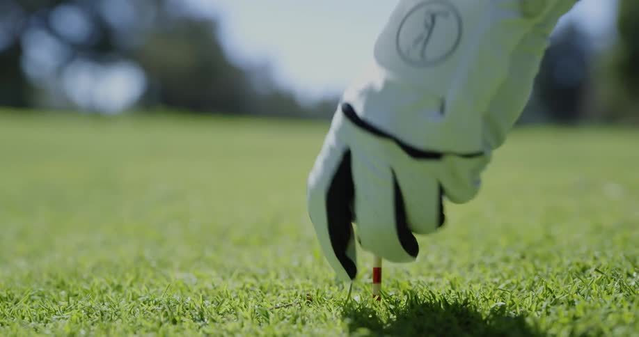 On a wide open grassy golf range a person places a golf tee into the ground with a white golf ball on top