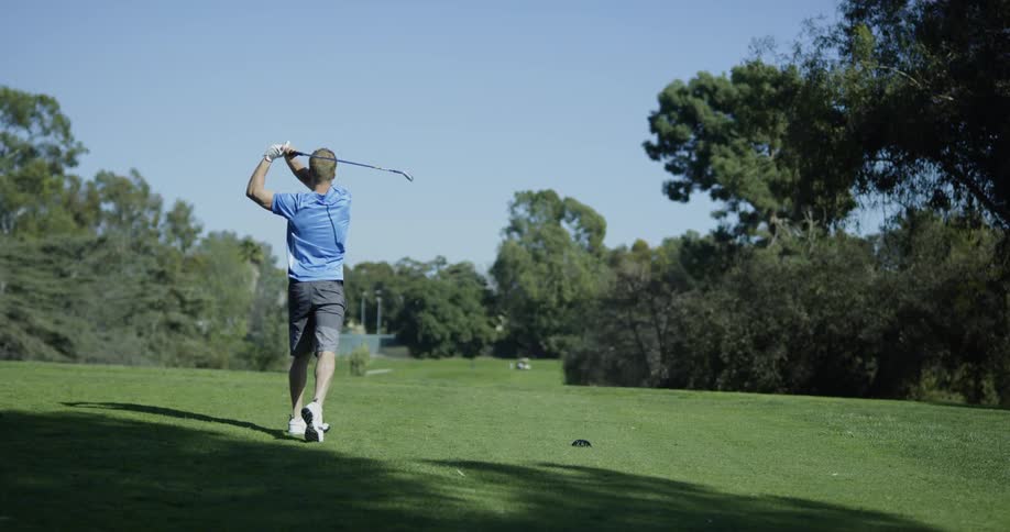 A man stands on a grassy green golf course preparing to hit the golf ball and then drives the ball off into the distance