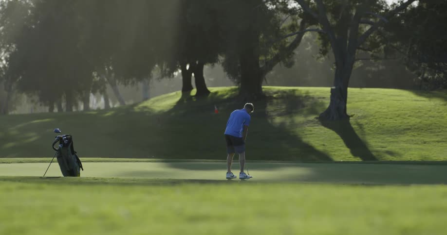 A white male crouches down to line up a shot on the green before approaching the golf ball and hitting it with his putter