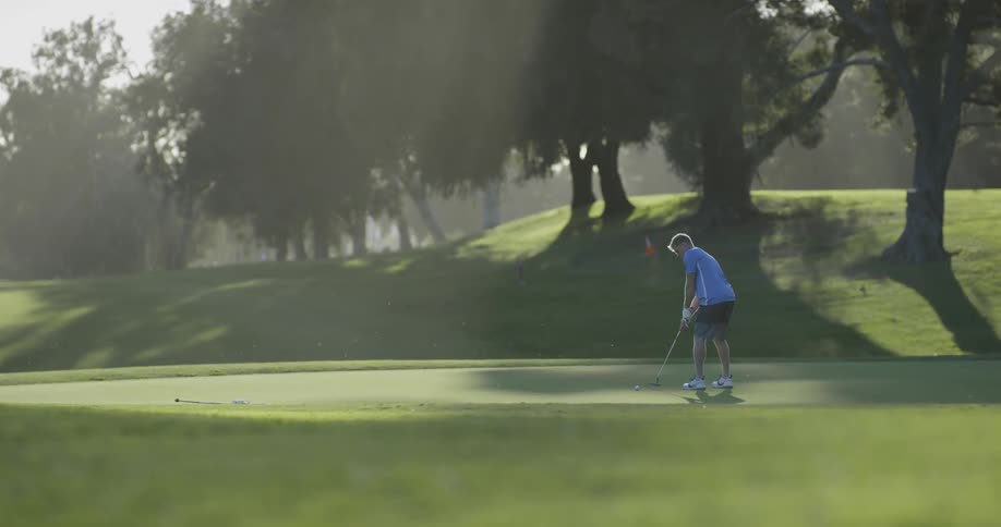 A white male golfer uses a putter to gently hit a golf ball into the whole on the putting green at a golf course