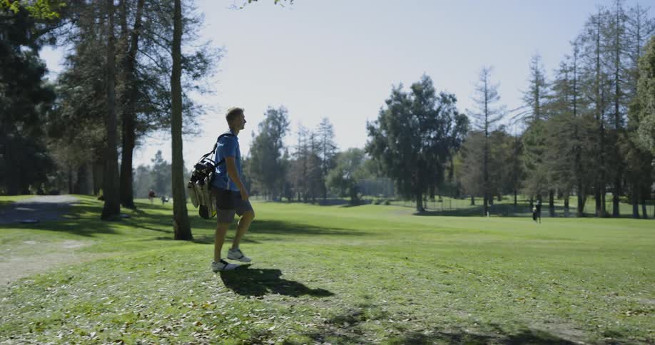 A man with golf clubs walks across a green course
