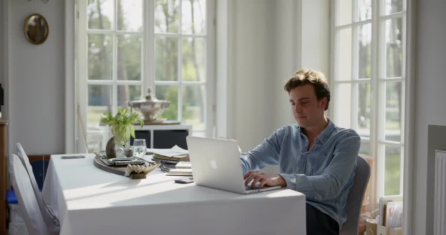 A man works on his laptop at a dining room table in front of two big windows