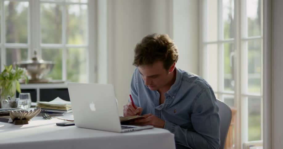 A man sitting at a large table types on his laptop and then jots something into a small notebook