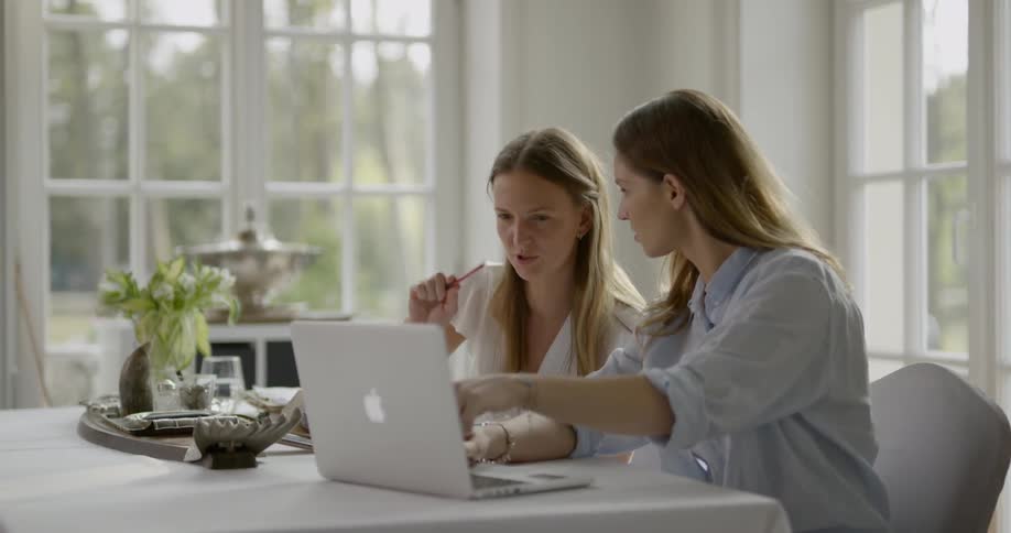 Two young women sitting at the table use laptop and write down on the notebook with a red pen in the room