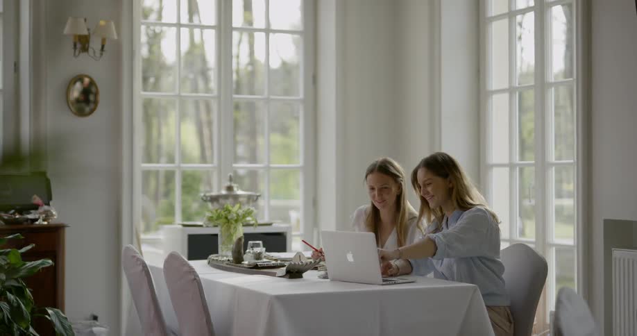 Two women sit working at a table one typing on a laptop and the other taking notes