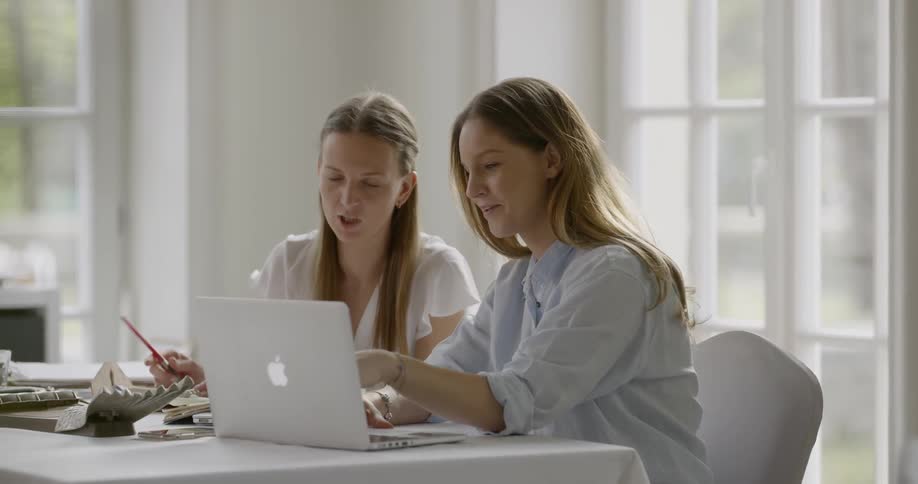 A young woman using a laptop while talking with a older woman who is writing in a notebook