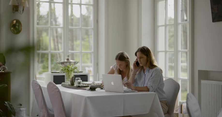 Two women sit at a table and write talk on the phone and work on a laptop