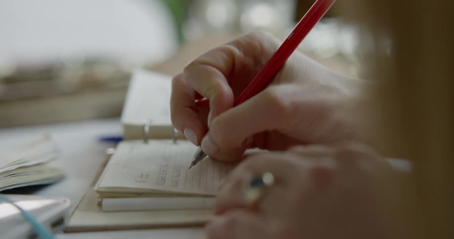 Woman sitting at the desk writes down in a small notebook with red pen in a room