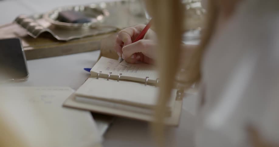 Blonde woman sitting at the table writes down on the notebook with a red pen in the room