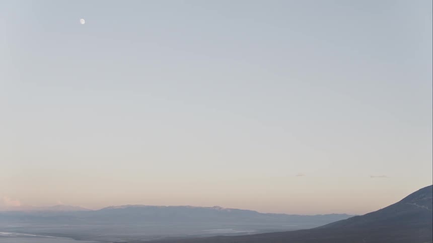 The moon is visible in the sky during daytime high above a range of mountains and the sprawling valley between them