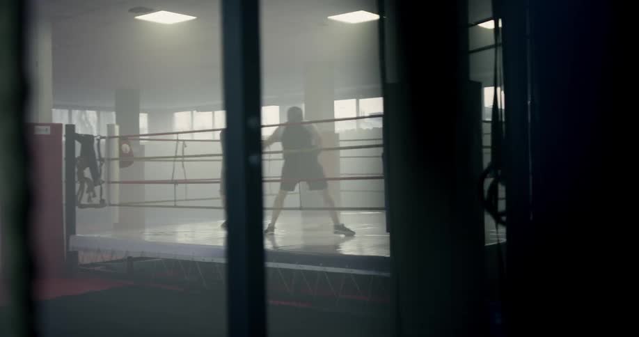 In a dimly lit gymnasium to men in a boxing ring spar while wearing gloves and protective headgear