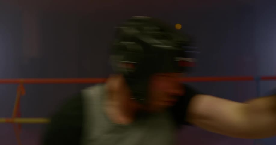 Lit only by a few spotlights two men in a boxing ring spar inside of a gym