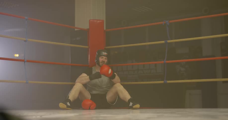 Male boxer sits in the corner of a boxing rings as he puts on his red and black boxing gloves