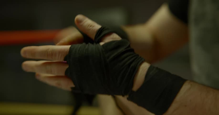 A man sitting in a boxing ring quickly undoes the black boxing wrap that is on his right hand