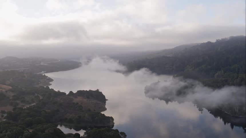 A large body of water sits between forest covered hills and mountains under very cloudy skies
