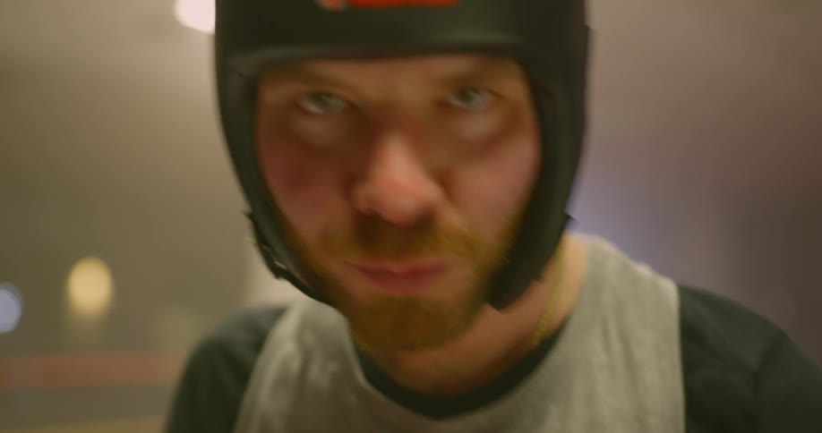 A man wearing boxing gloves and padded headgear has a look of determination on his face as he prepares to spar in a boxing ring