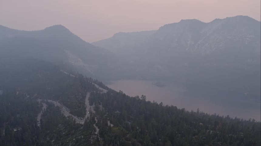 A curving road cuts through the forest trees above a mountain lake at sunset