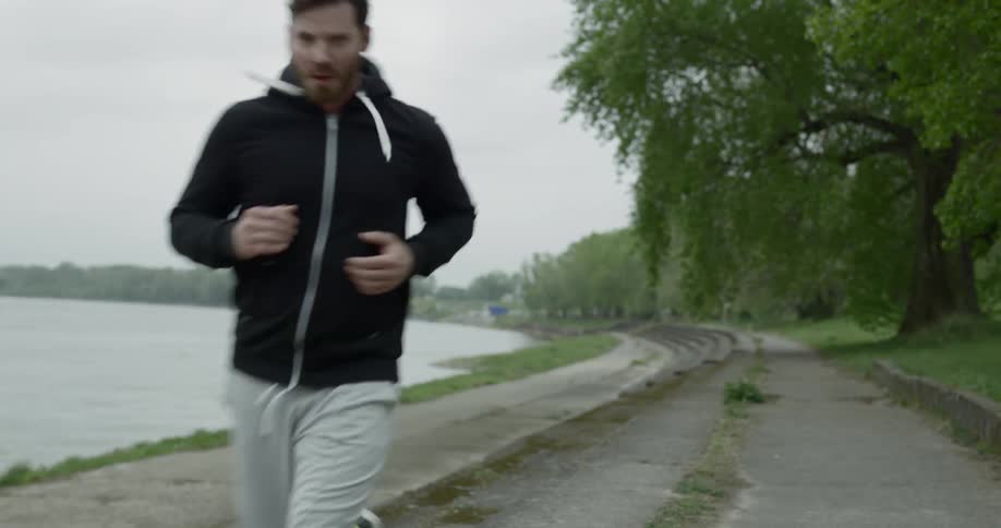 A white male in a black hooded sweatshirt runs on a paved path next to a lake