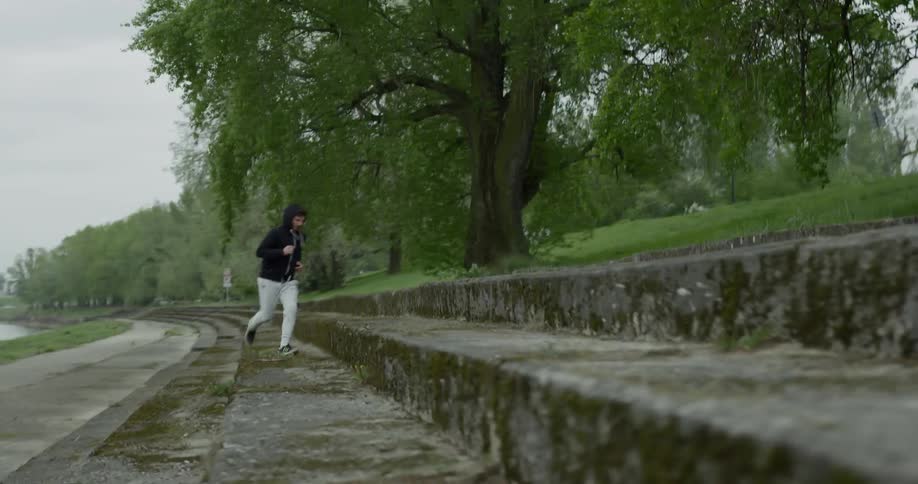 A man is jogging on a paved path next to a body of water and then runs up a few cement steps to continue jogging on another paved path