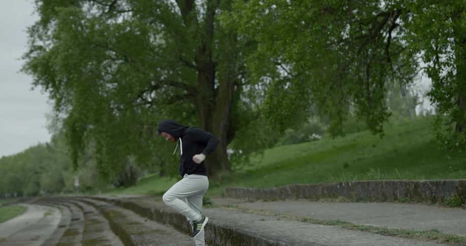 A young male stands outside on some concrete steps near a grove of trees as he practices boxing maneuvers