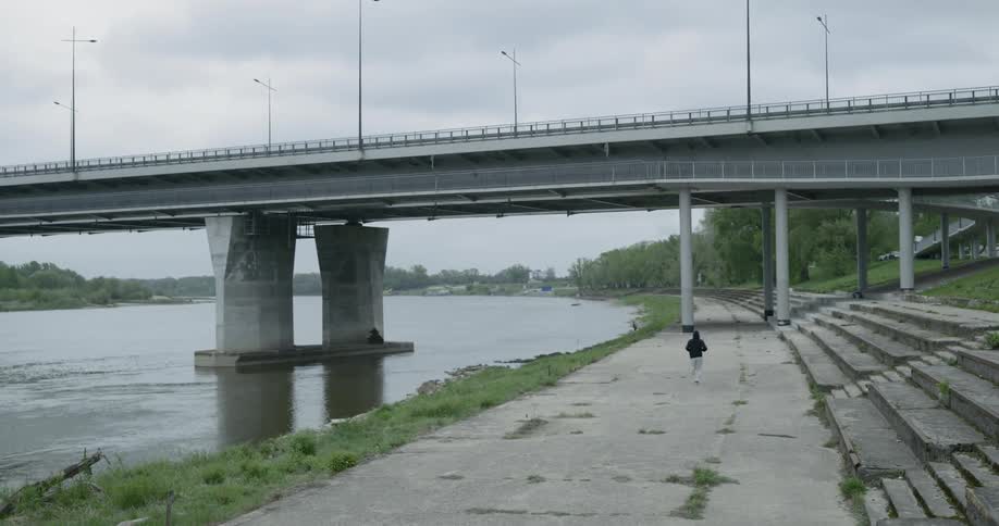 Man runs along the road next to the river under the bridge on a cloudy day