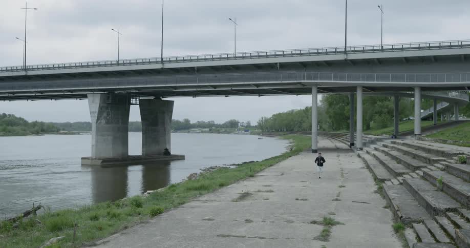 A young male runs along a concrete path on the bank of a long river that passes under a bridge