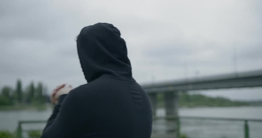 A man wearing a hoodie stands outdoors near a river as he warms up before exercising