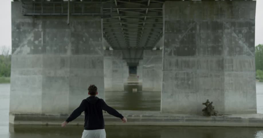 Man in black hoodie warms up for running next to the river under the bridge