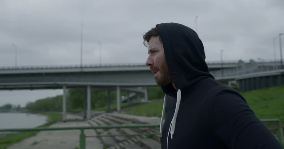 A man wearing a hoodie stands outdoors near a river and a bridge on a cloudy day