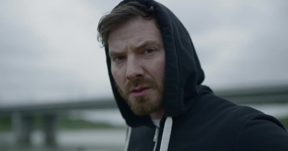 A man with a beard and hoodie staring with a bridge in the background