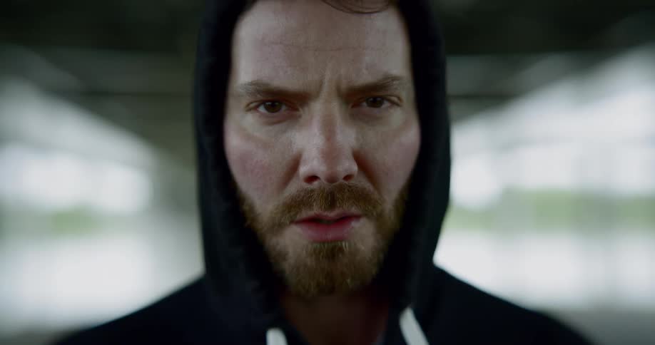 Bearded man wearing a black hoodie raises his head and looks at the camera angrily in the building