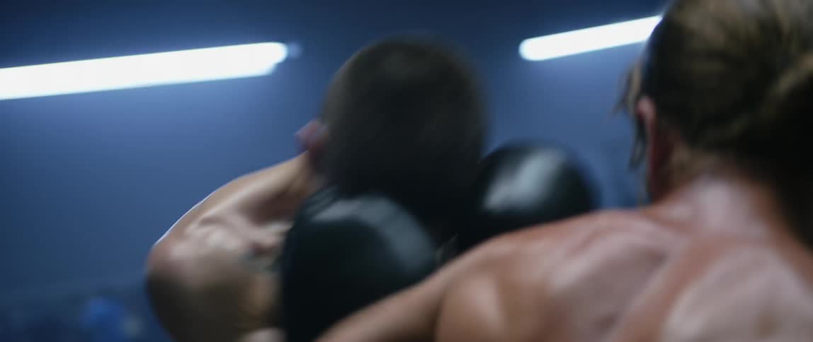 Two men boxing shirtless in a boxing ring under fluorescent lights