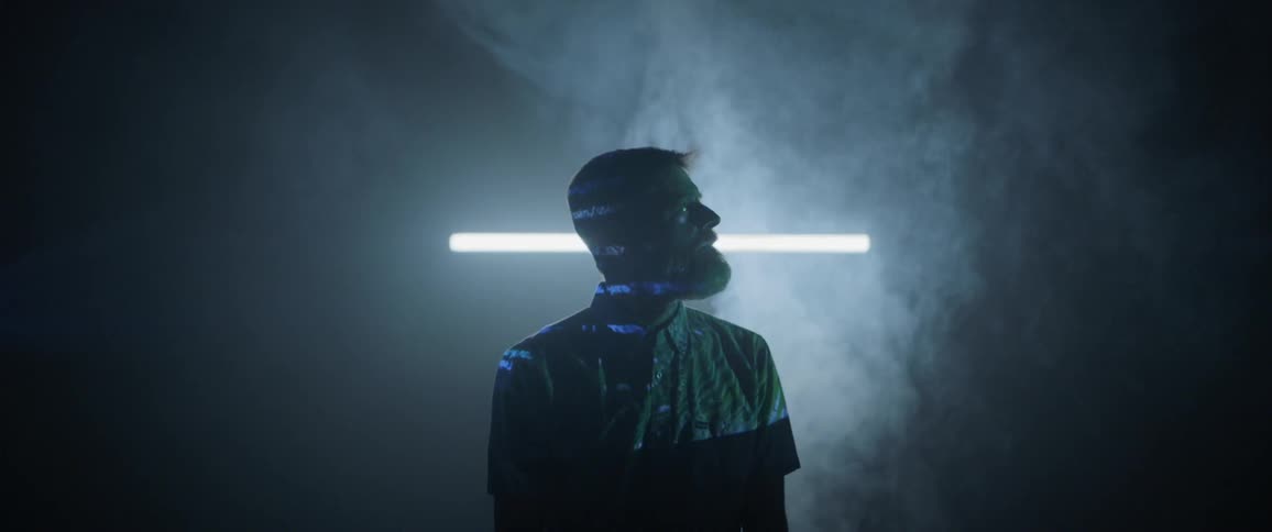 A man with a large beard stands in a dark room with fog wafting upwards behind him as computerized text is projected onto his face