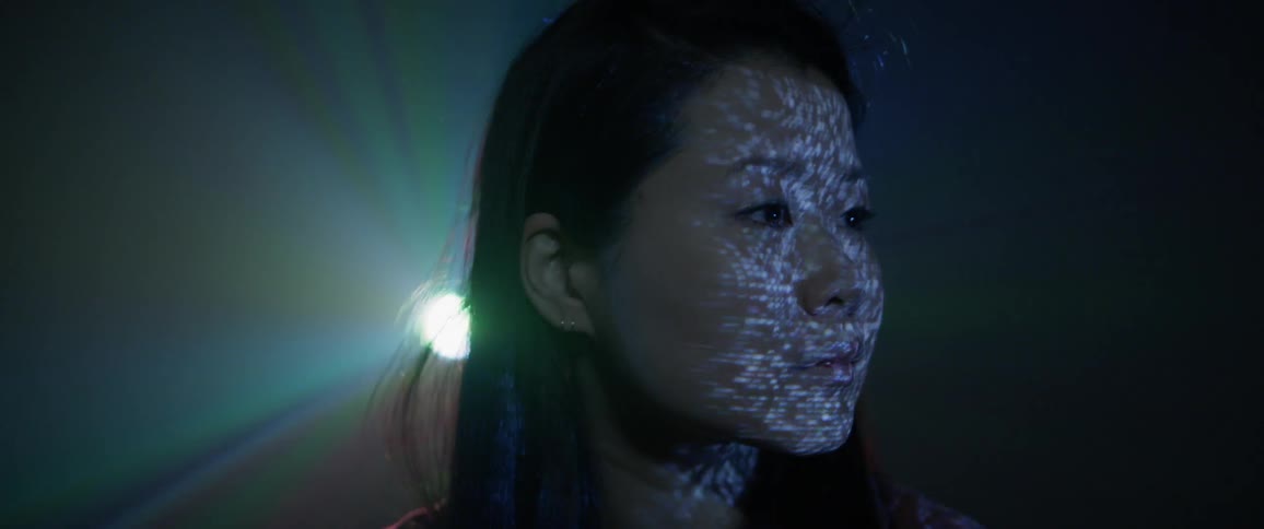 A woman stands in a dark room with lighted designs projecting on her skin