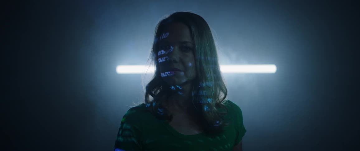A woman stands in a dark and hazy room as lines of computerized text are projected onto her face