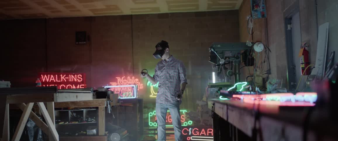 A man holding controllers and wearing a vr headset stands in a room full of neon signs and work benches