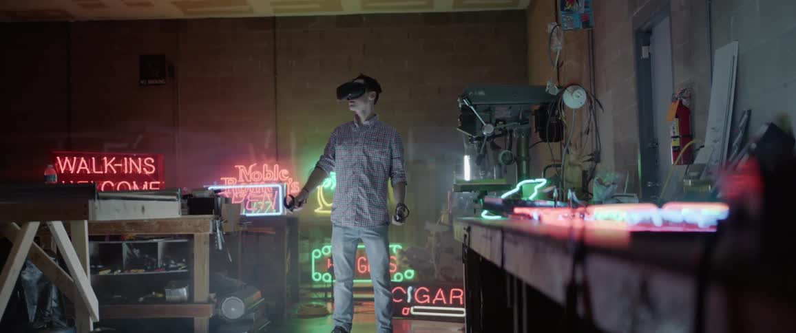 A man wearing a virtual reality headset stands in the middle of a work room surrounded by neon signs