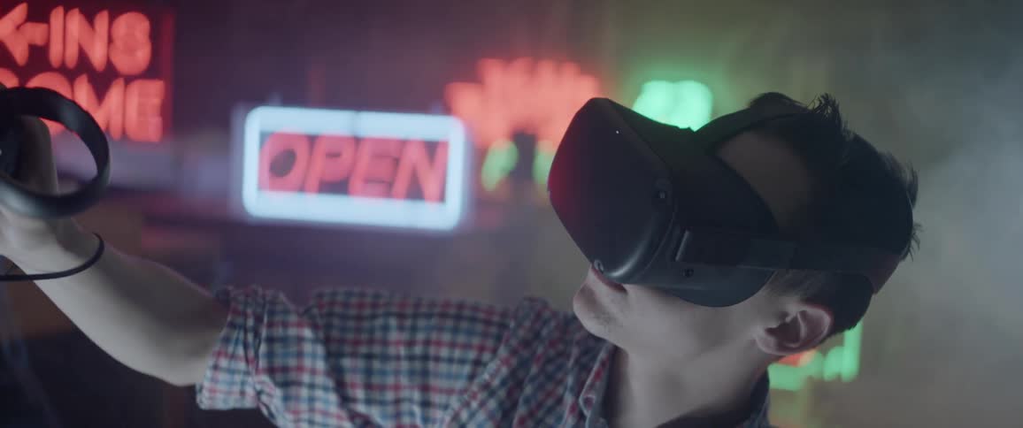 A man wearing a vr headset moves a controller around in a room full of neon signs