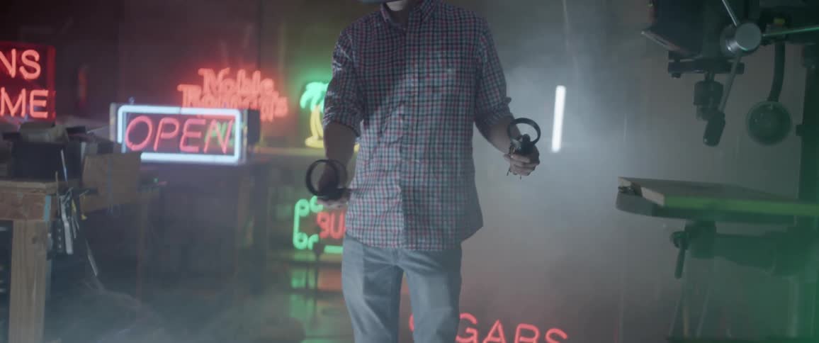 A man stands in a hazy room with numerous neon signs while he waves around controllers while wearing a virtual reality headset