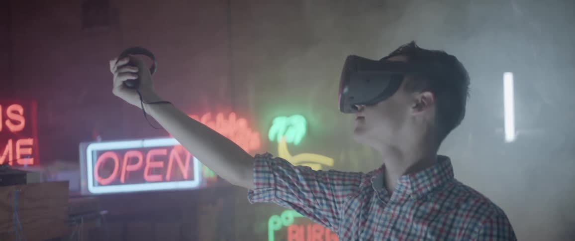 A young male holds out a controller in his hand while wearing a virtual reality headset in a smokey room filled with neon signs