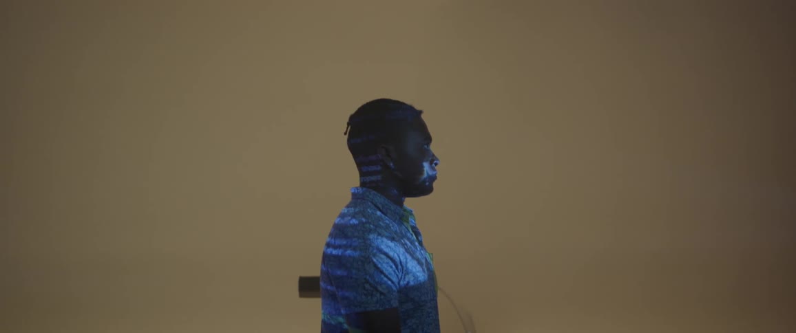 A man stands in an empty room while words are projected onto his face and body