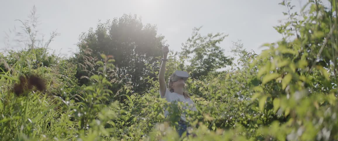 A girl stands amidst plants and shrubs while using vr glasses