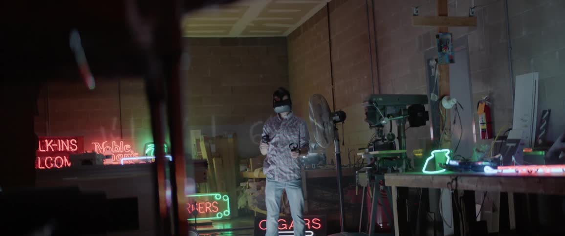 A man holds two objects as he looks through a virtual reality headset in a warehouse with tools and neon signs