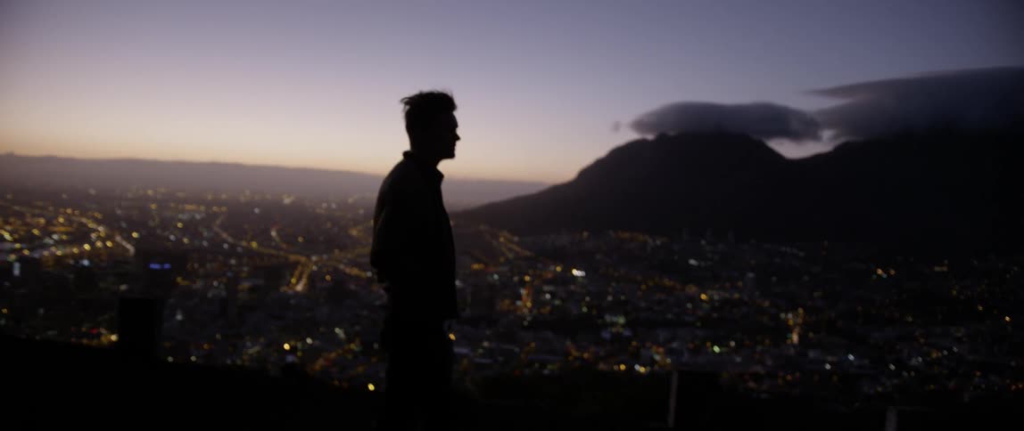 A young man stands on top of a hill and looks down at the city lights