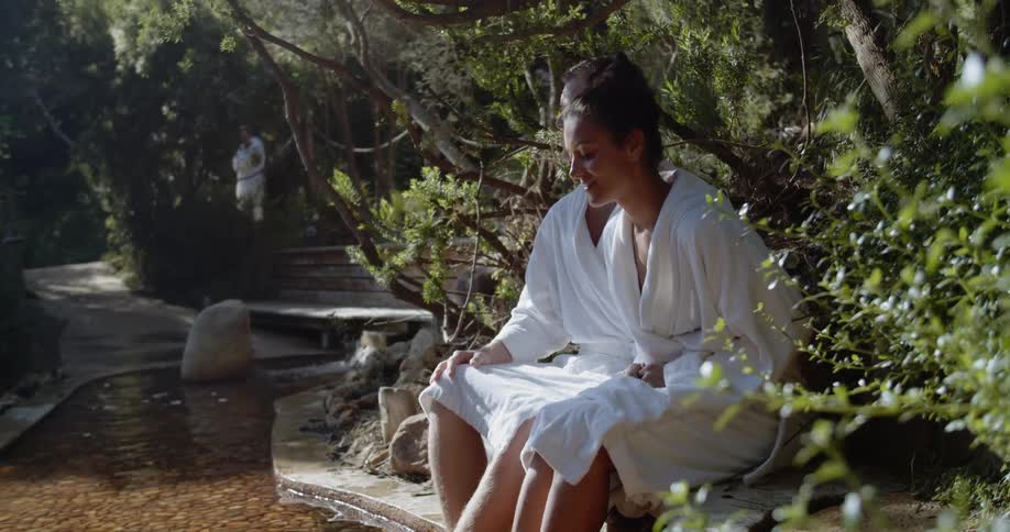A man and woman sit with their feet in a creek while wearing robes